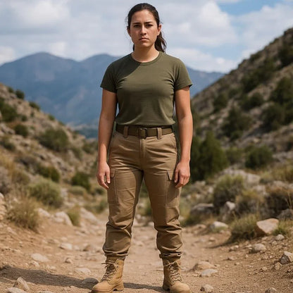 Femme en tenue tactique portant des bottes style rangers beiges, debout sur un sentier de montagne entouré de végétation et de sommets